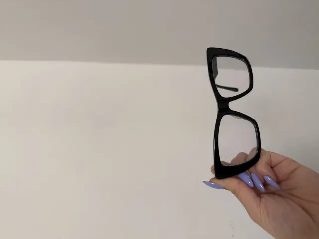 A phone photo: black-rimmed clear glasses held in a hand against a plain wall